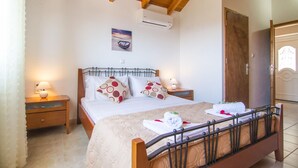 Un dormitorio con techo de madera, una cama con cubrecama color beige, dos almohadas estampadas con lunares, una mesita de noche con lámpara y un equipo de aire acondicionado en la pared.