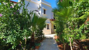 A pathway leading to a white building with balconies, surrounded by lush greenery and flowering plants. - Villa Eos in Kalkan with 3 bedrooms and 4 bathrooms (Kalkan)