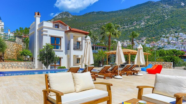 A poolside area with lounge chairs and umbrellas, a pool, and a view of a hillside town. - Villa Eos in Kalkan with 3 bedrooms and 4 bathrooms (Kalkan)