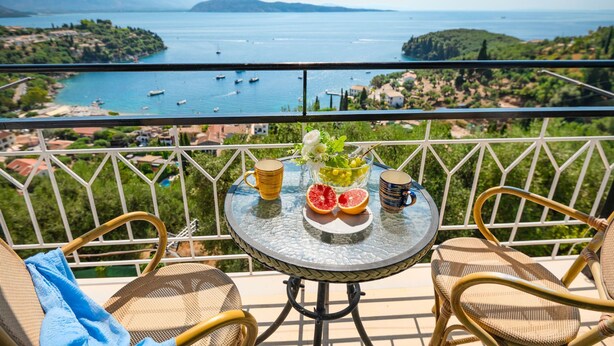 Un balcón con sillas de mimbre, una mesa con frutero y vistas al mar y las colinas.