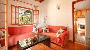 Una sala de estar acogedora con techo de madera, un sofá rojo con cojines estampados, una mesa de centro de vidrio y vistas al exterior a través de una ventana con persianas de madera.