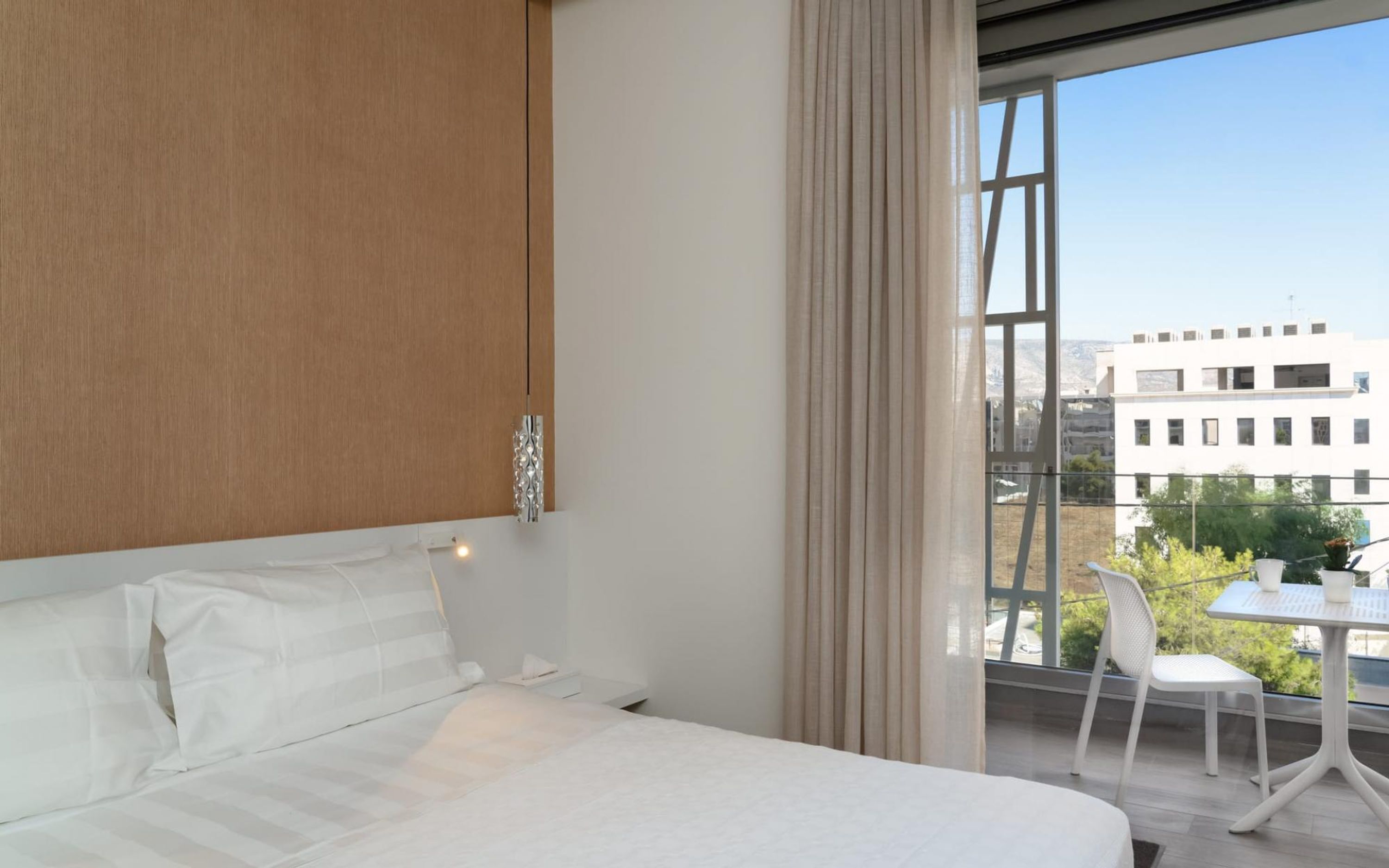 A modern bedroom with a large bed, a wooden headboard, a hanging light fixture, and a view of buildings through a large window.