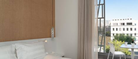 A modern bedroom with a large bed, a wooden headboard, a hanging light fixture, and a view of buildings through a large window.