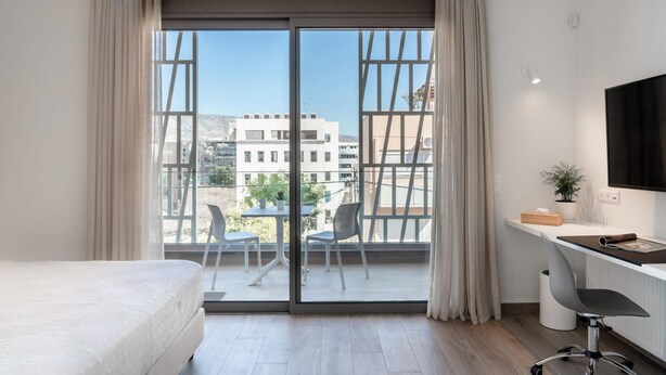 A modern hotel room with a large sliding glass door to a balcony, a bed, a desk with a chair, and a TV on the wall. - Athens Hill - Superior Apartment with Balcony (Kallithea)