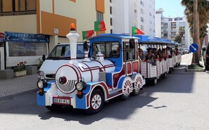 A colorful train with passengers, parked on a street with buildings and palm trees in the background. - T0 A/C Varandas Mar 3ºB perto PRAIA Wi-Fi (Quarteira)