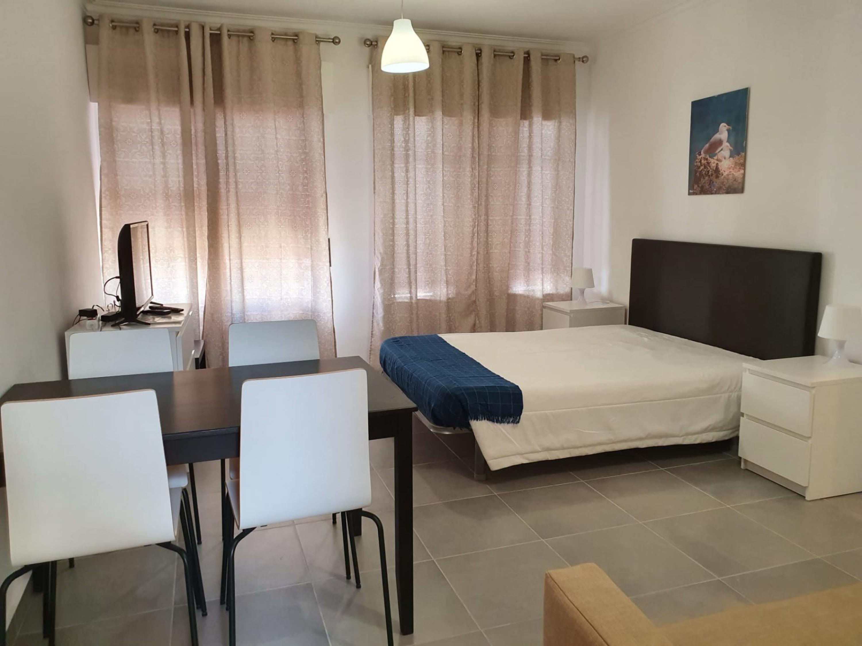 A hotel room with a bed, a desk with a computer, a chair, a bedside table with a lamp, and a painting on the wall.