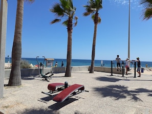 A beachside exercise area with stationary bikes, a red lounge chair, and palm trees. - T0 A/C Varandas Mar 3ºB perto PRAIA Wi-Fi (Quarteira)