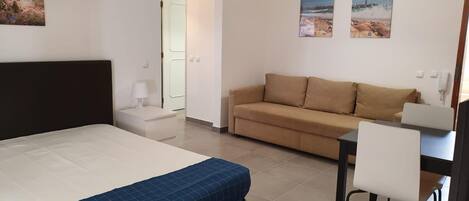 A hotel room with a bed, a sofa, a desk with a chair, and two paintings on the wall.