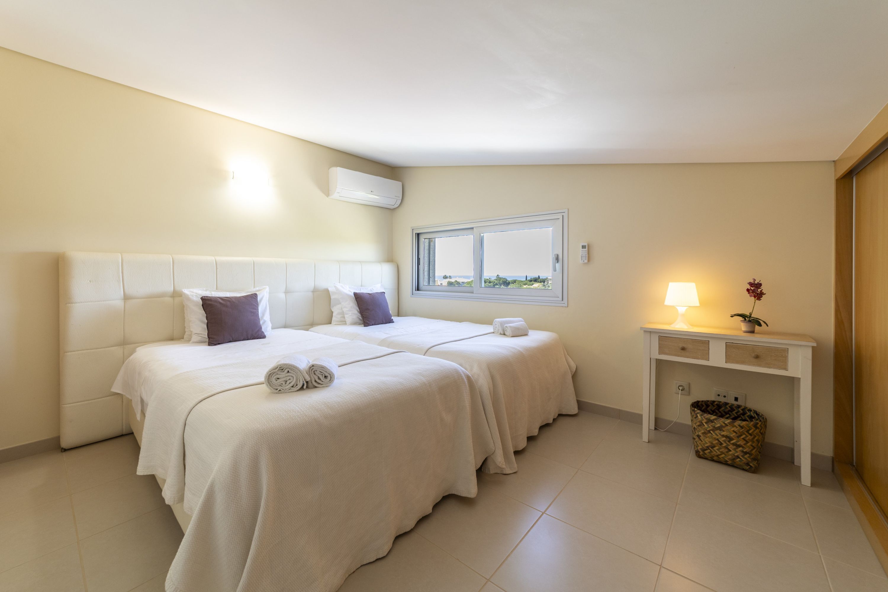 A hotel room with two beds, a bedside table with a lamp, a window, and a wall-mounted air conditioner.