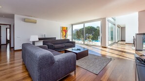 Ein modernes Wohnzimmer mit einer Couch, einem Couchtisch und einem Fernseher. Ein großes Fenster bietet Blick auf Bäume und einen Pool.
