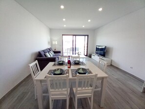 A modern living room with a dining table set for two, a sofa, a TV, and a large window. - T3 Alto Forte -Vista Mar,Ar Condicionado,Garagem (Quarteira)