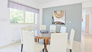 A modern dining room with a round table, white chairs, a large window with a view of greenery, and a colorful abstract painting on the wall. - Villa Georgios Rafaela (Skoulli)