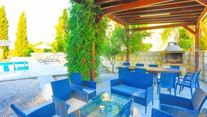 An outdoor patio with wicker furniture, a glass table, and a stone wall. - Villa Georgios Rafaela (Skoulli)