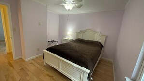 Quarto com cama branca, ventoinha no teto, mesa de cabeceira com abajur e janela.