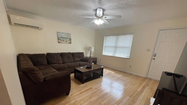 Uma sala de estar com sofá seccional, mesa de centro, ventilador de teto, um quadro e uma janela com persianas.
