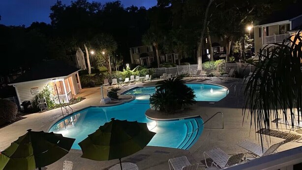 A swimming pool with multiple sections, surrounded by lounge chairs and umbrellas, and a small building in the background. - Newly Remodeled Family and Pet Friendly Condo townhouse  in Ocean Walk (St. Simons Island)