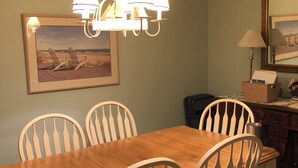 A dining area with a wooden table and white chairs, a chandelier, a framed picture on the wall, and a desk with a lamp and a box. - Newly Remodeled Family and Pet Friendly Condo townhouse  in Ocean Walk (St. Simons Island)