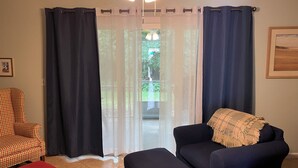 A living room with a checkered armchair, a dark sofa with a throw blanket, a red ottoman, and a view of a garden through sheer curtains. - Newly Remodeled Family and Pet Friendly Condo townhouse  in Ocean Walk (St. Simons Island)