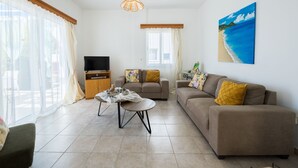 A living room with a sofa, a coffee table, a television, and a painting of a beach scene. - Lemon Grove Villa Ena (Argaka)