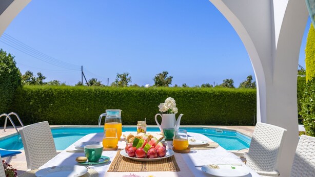 A dining table with a view of a swimming pool, a plate of fruit, and a pitcher of orange juice. - Lemon Grove Villa Ena (Argaka)