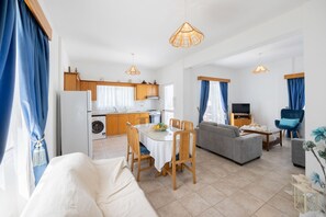 A modern living room with a sofa, dining table, and chairs, a kitchen with wooden cabinets, and a TV. - Lemon Grove Villa Dio (Argaka)