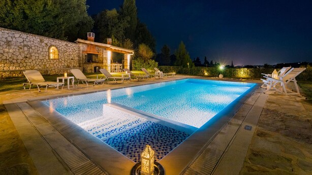 Una piscina rettangolare con muro in pietra e sedie a sdraio intorno, circondata da alberi e un edificio in pietra.