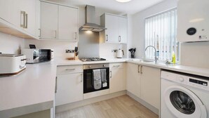A modern kitchen with white cabinetry, a built-in oven, a microwave, a washing machine, and a window with blinds. - Modern 3 Bed Home with Garden, Free Parking, Wi-Fi (Lazenby)