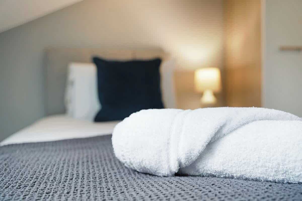 A neatly made bed with a white towel, a grey textured bedspread, and a headboard with a dark pillow.