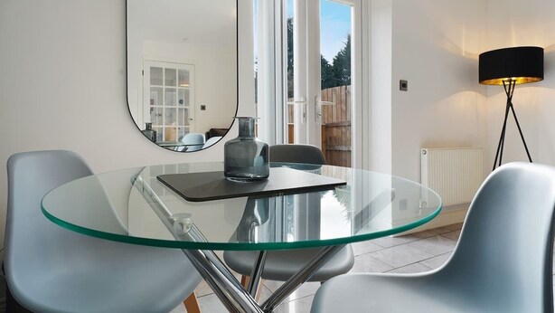 A modern dining area with a glass table, two chairs, a mirror, and a floor lamp. - Modern 3 Bed Home with Garden, Free Parking, Wi-Fi (Lazenby)