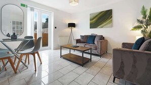 A modern living room with a glass dining table, a sofa, a coffee table, a floor lamp, and a large mirror. - Modern 3 Bed Home with Garden, Free Parking, Wi-Fi (Lazenby)
