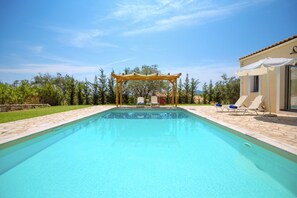 Una piscina con pérgola, sillas de descanso y un edificio de techo con tejas.