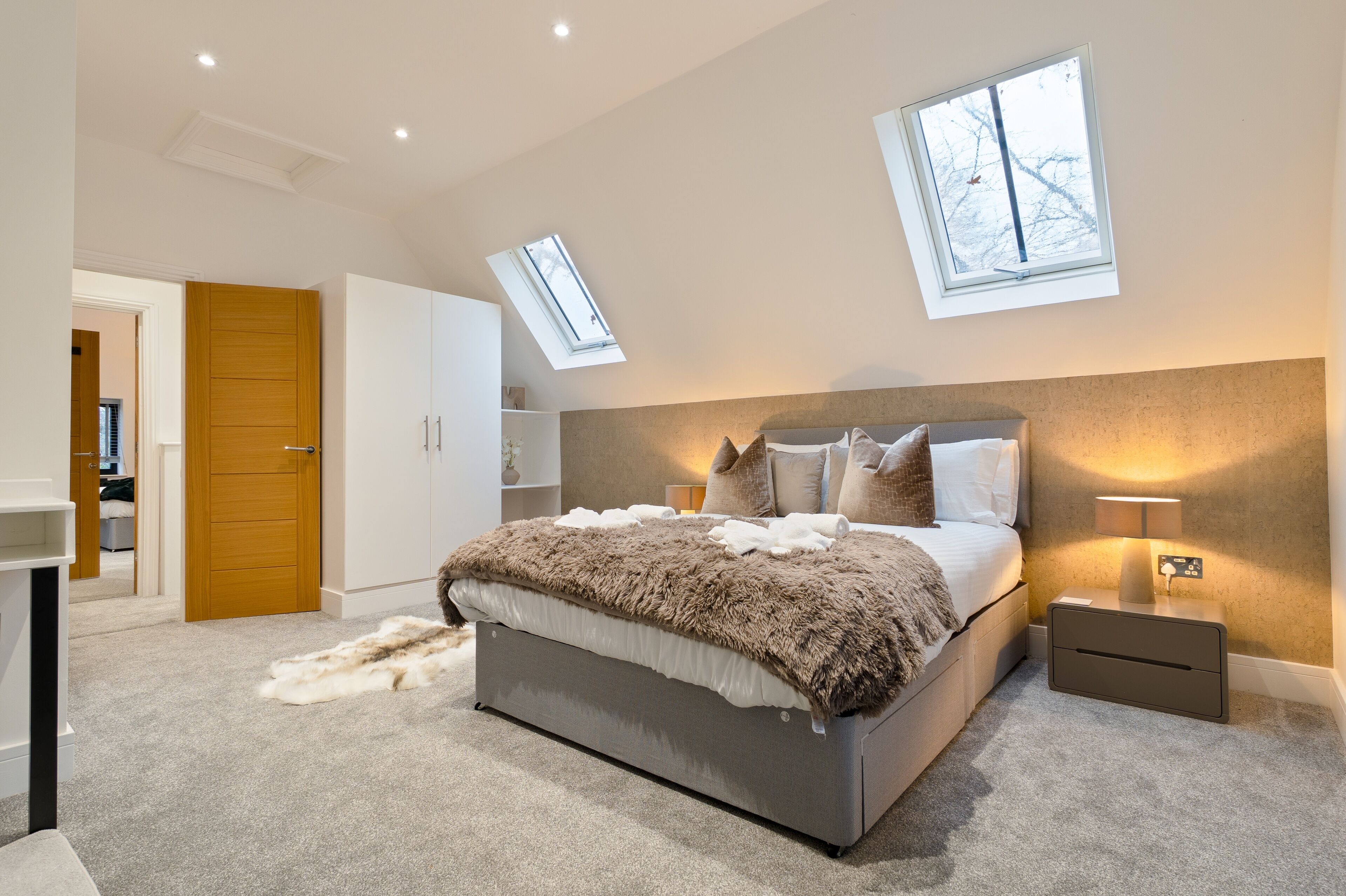 A bedroom with a large bed, a nightstand, a lamp, a wardrobe, and a skylight.