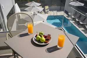 A table with a plate of fruit and chocolate, two glasses of orange juice, and a poolside setting with umbrellas and lounge chairs. - Athina Suites | A 2BD chic apartment w/shared pool (Platanias)