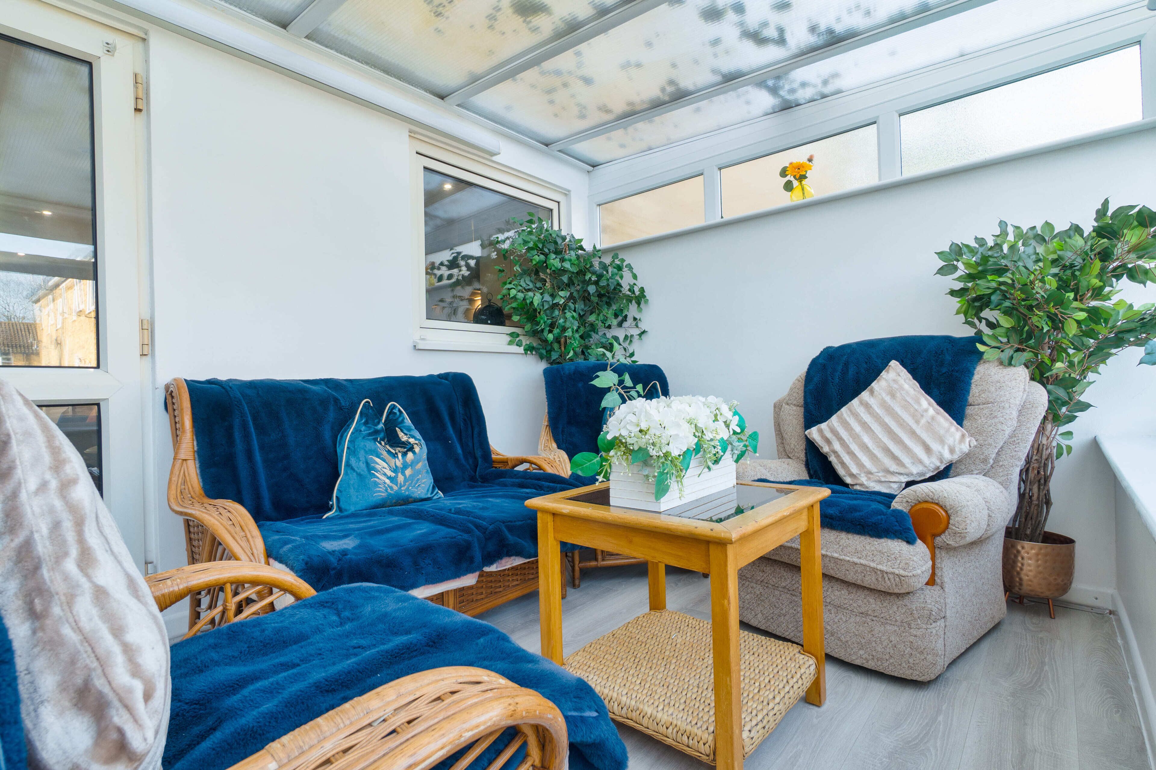 Une véranda meublée avec des meubles en osier, un canapé bleu, un fauteuil rembourré et des plantes en pot.