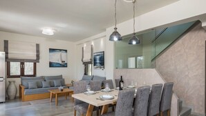 A modern living room with a sofa, dining table, and chairs. There is a staircase with a glass railing and a painting on the wall.