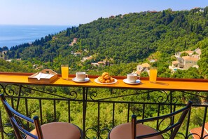 Ein Balkon mit Frühstückstisch, mit Blick auf einen üppigen, grünen Hang und das Meer.