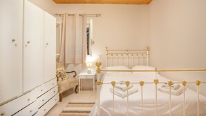 Een slaapkamer met een houten plafond, een wit bed met gouden accenten, een kledingkast, een nachtkastje met een lamp en een stoel met een bloemenkussen.