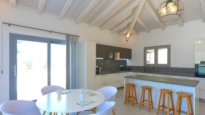 A modern kitchen with white cabinets, a large island with stools, and a dining area with a round table and chairs. - Villa Loki in Kremithas (Kremithas)