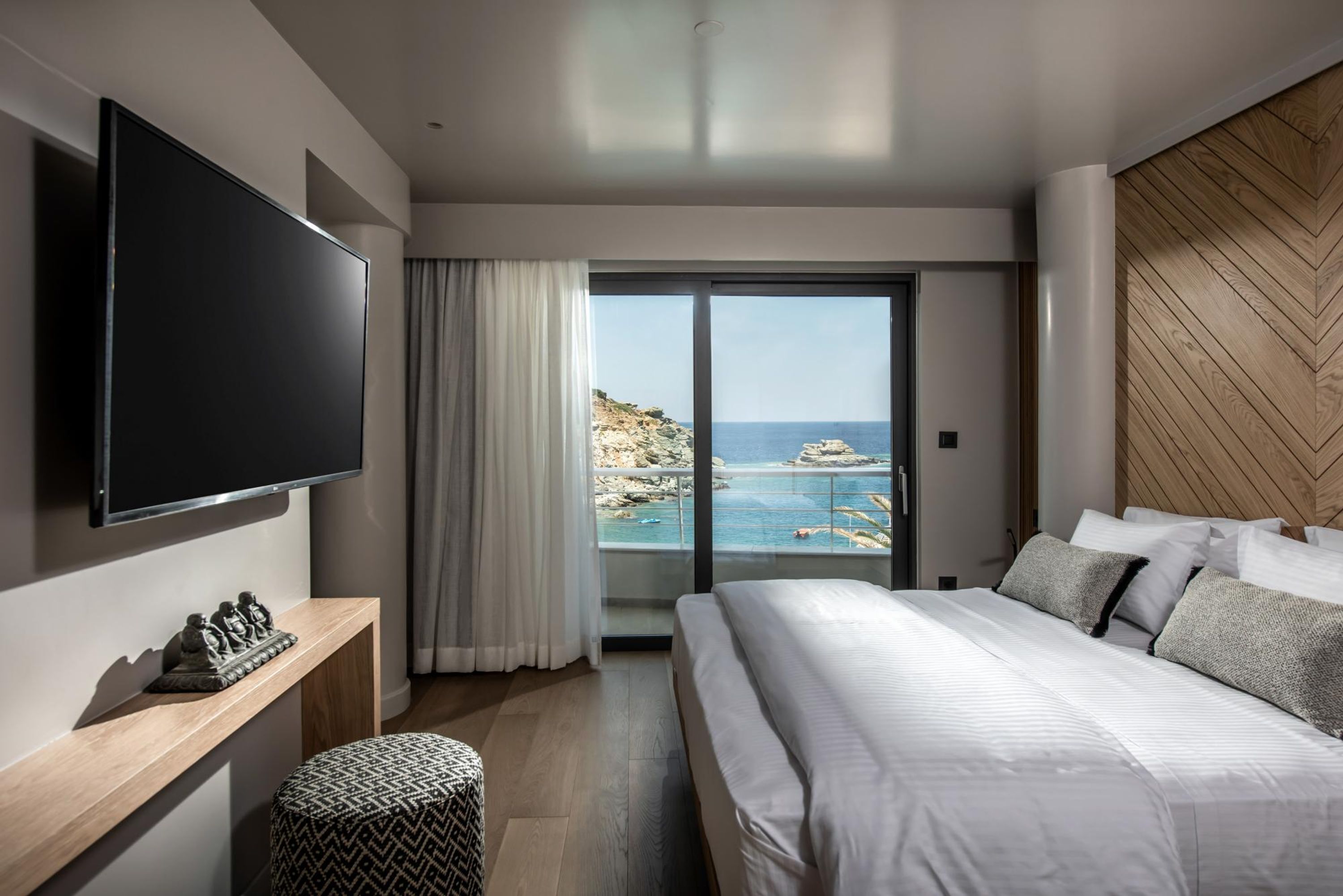 A modern hotel room with a large bed, a flat-screen TV, a wooden console table, and a view of the sea through a sliding glass door.