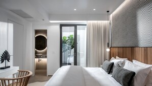 A modern bedroom with a large bed, a wooden chair, a bathroom with a round mirror, and a patterned wall. - Made By The Sea Villa 4 (Mades)