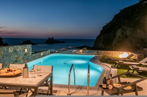 Área de piscina com vista para o oceano, cadeiras de descanso e uma mesa com garrafa de vinho e uma cesta de frutas.