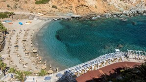 Vue aérienne d’une plage aux eaux turquoise et limpides, bordée d’une côte rocheuse, et où l’on aperçoit une série de transats et de parasols disposés le long du rivage.
