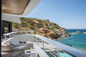 A balcony with white furniture and a table, overlooking a rocky coastline and a clear blue sea. - Made By The Sea Villa 2 (Mades)