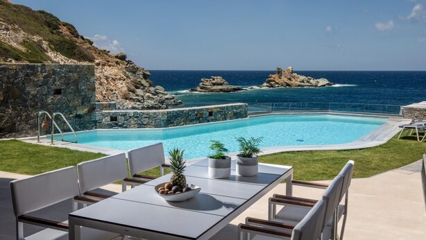 Une piscine avec vue sur la mer et les falaises rocheuses, une table avec des chaises et des plantes en pot.