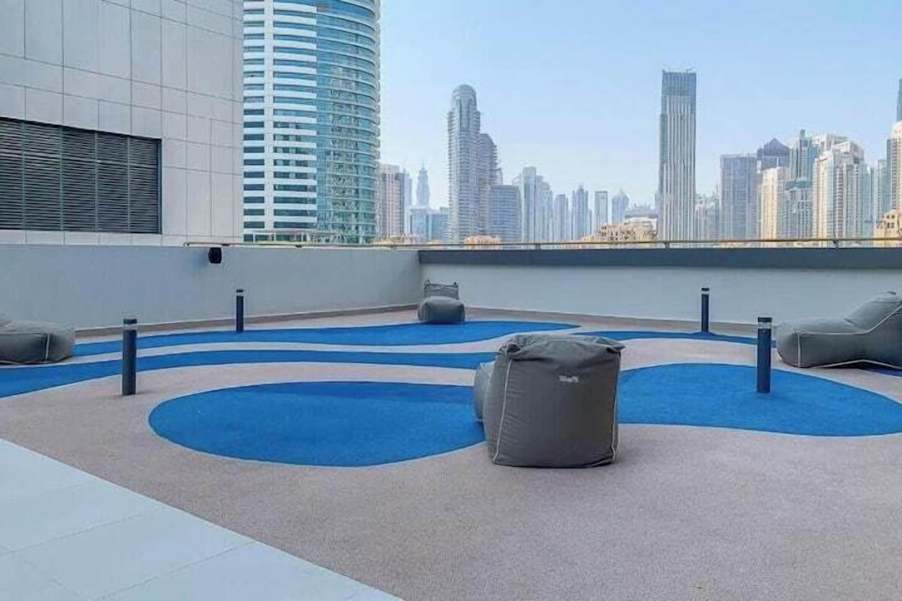 A rooftop terrace with blue circular carpet patterns, grey bean bags, and a view of a city skyline.