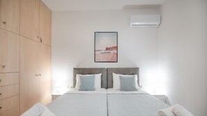 Een tweepersoonsbed met twee kussens, een houten garderobe, een lijst met een foto aan de muur en een opgerolde handdoek op het bed.