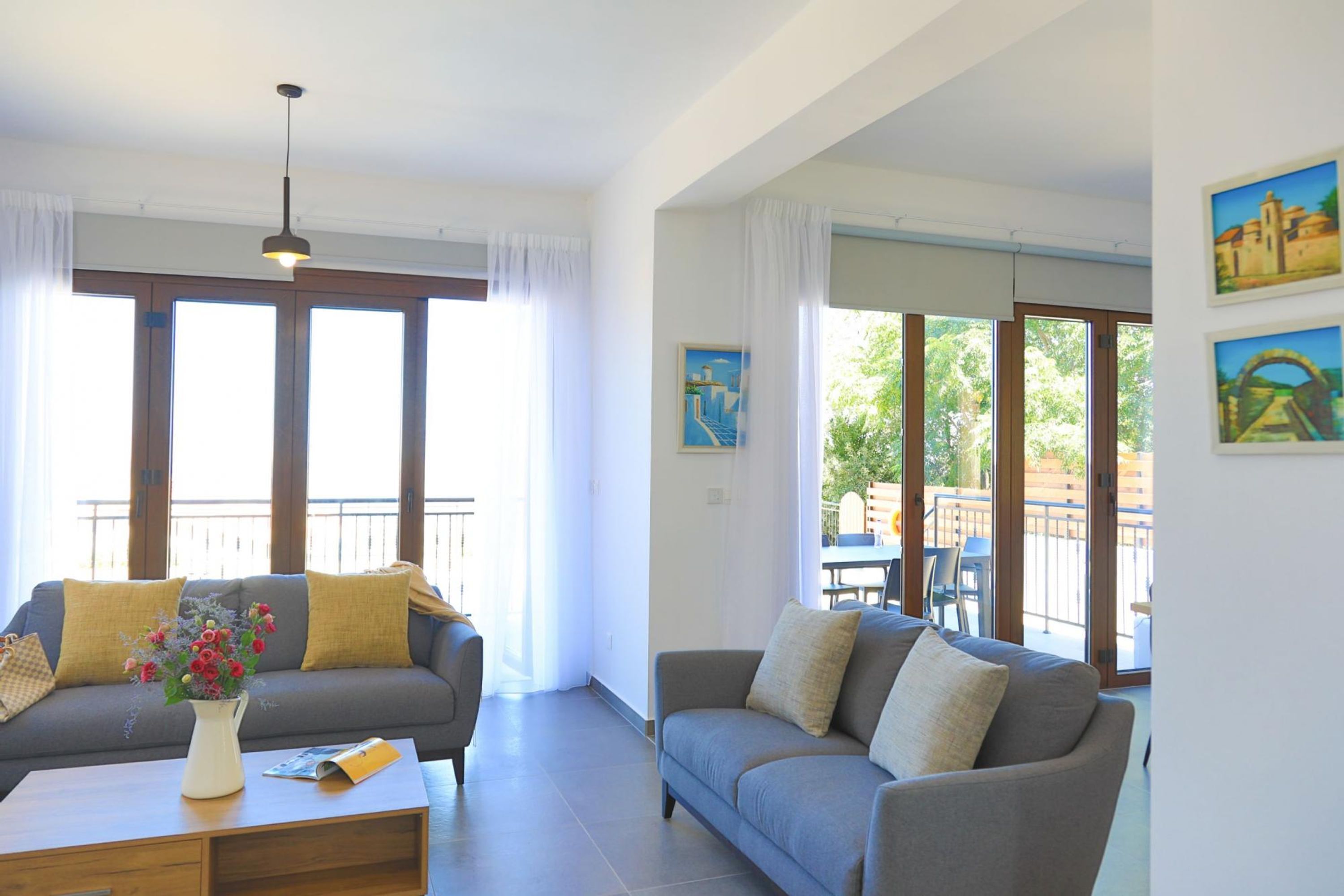 A living room with two sofas, a coffee table, and a vase of flowers. There are large windows with curtains and a balcony visible through the windows.