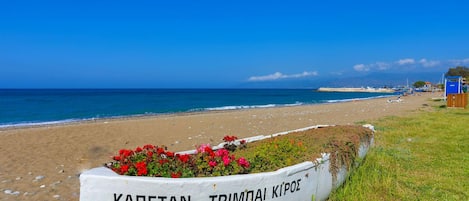 Ein Strand mit einem weißen Boot mit der Aufschrift "ΚΑΠΕΤΑΝ ΤΡΙΜΠΑΛΑ ΚΙΡΟΣ" und einer kleinen blauen Struktur im Hintergrund.