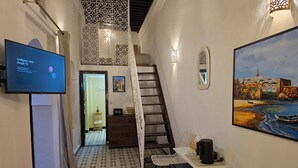 Una camera d'albergo con una scala, una TV che mostra l'interfaccia Google TV, un quadro raffigurante una veduta costiera e un tappeto con motivo decorativo.