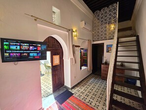 A hallway with a TV displaying a comedy channel, a wooden door with a sign, a patterned floor, and a staircase. - Riad Boutique Baba (Rabat)